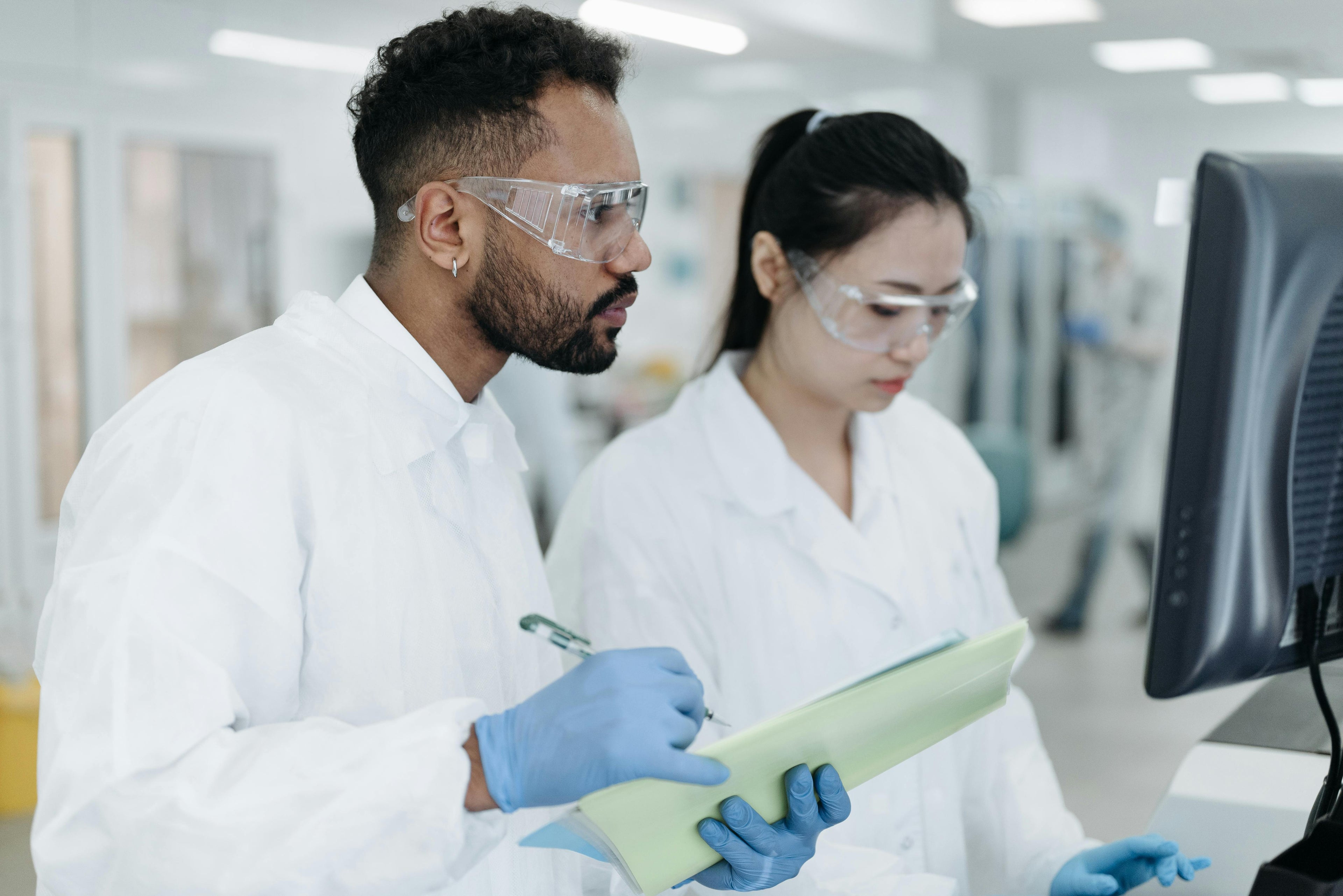 Two scientists in a lab setting, one holding a document and the other looking at a computer screen.