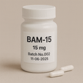 White plastic pill bottle labeled "BAM-15, 15 mg, Batch No.002, 11-06-2025" standing on a beige background, with two white capsules placed beside it.