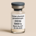 A digital photograph of a transparent glass vial with a gray metal cap, labeled "Human Chorionic Gonadotropin (HCG), 5000 IU, Batch No.007, 20-09-2025." The vial contains a fine white powder at the bottom and stands upright on a beige surface against a minimalist background. The clean white label with bold black typography emphasizes the professional pharmaceutical design.