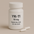 White plastic pill bottle labeled “YK-11 10 mg — Batch No. 004 — 05-08-2025” with two white capsules beside it on a neutral beige background.