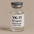 A transparent glass vial filled with clear liquid, labeled ‘YK-11 70 mg/mL – Batch No.002 – 17-07-2025,’ sealed with a gray rubber stopper and matte aluminum cap, set against a soft beige background